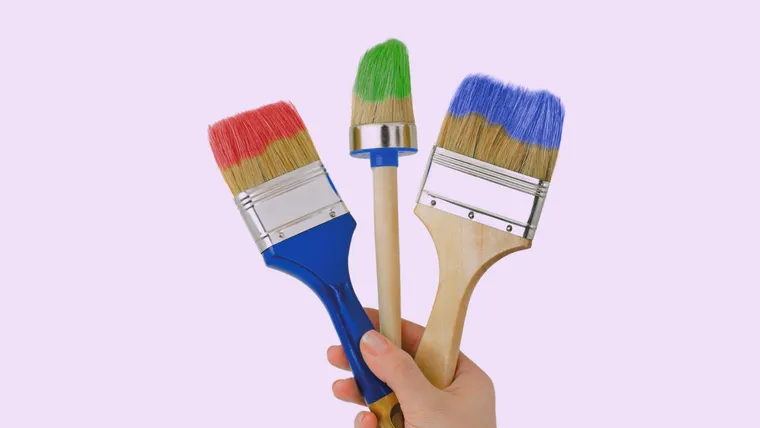 Eine Hand hält drei unterschiedliche Pinsel mit Farben in die Höhe.