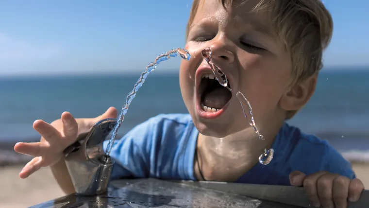 Einem kleinen Kind am Strand spritzt Wasser aus einem Wasserhahn in den Mund